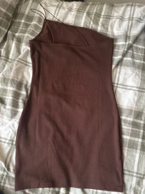 Brun ribbad kortklänning från DIVIDED - Säljer en brun ribbad kortklänning från DIVIDED i storlek M. Klänningen har smala axelband och asymmetrisk halsringning som ger en cool look. Materialet är mjukt och stretchigt, perfekt för dig som gillar stilrena och enkla plagg.