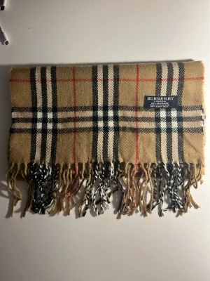 Rutig beige Burberry väst i kashmir - Säljer en klassisk väst från Burberry i 100% kashmir. Västen har det ikoniska rutiga mönstret i beige, svart, vitt och röda linjer. Den är mjuk och har fransar längst ner. Perfekt för dig som gillar lyxiga och tidlösa plagg.