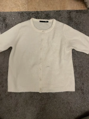 Vit kofta från 157, storlek XS - Mysig vit kofta från 157 i storlek XS. Koftan har rund halsringning och knäpps med sju vita knappar framtill. Materialet är stickat och känns mjukt mot huden, perfekt för lager-på-lager under kyliga dagar.
