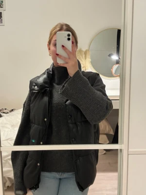 Svart dunväst i skinnimitation - Säljer en svart dunväst i skinnimitation. Västen har hög krage, dragkedja och tryckknappar framtill samt två fickor. Perfekt att slänga över en stickad tröja för en cool och trendig look.