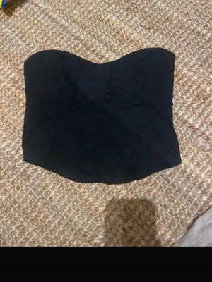 Svart bandeau tubtopp från Gina Tricot - Säljer en svart bandeau tubtopp från Gina Tricot i storlek S. Toppen är axelbandslös och har en stilren, enkel design med lätt formad överdel. Materialet är mjukt och stretchigt, perfekt för att skapa en snygg siluett. Passar dig som gillar minimalistisk stil.