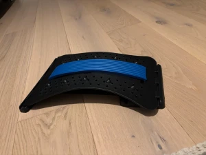  ryggsträckare - Säljer en ergonomisk ryggsträckare i svart plast med blått gummiband i mitten. Den har en böjd form och är täckt med små massagepunkter för extra effekt. Perfekt för att stretcha ryggen hemma eller efter träning.