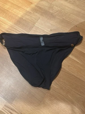 Svart bikiniunderdel från Lindex - Svart bikiniunderdel från Lindex i storlek 46. Klassisk trekantsform med enkel design och en liten metallring på sidan. Materialet är mjukt och stretchigt, perfekt för strand eller pool.