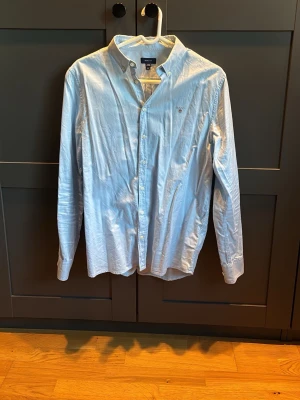 Ljusblå skjorta från GANT 170cm - Snygg ljusblå skjorta från GANT med klassisk krage och knappar hela vägen. Skjortan har lång ärm och diskret logga broderad på bröstet. Tillverkad i mjuk bomull som känns skön mot huden. Perfekt för dig som gillar stilrena plagg med enkel design.