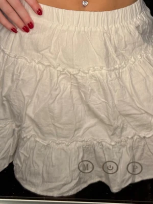 Vit volangkjol med resårmidja - Supersöt vit kortkjol med flera volanger och resår i midjan. Kjolen är gjord i mjukt bomullstyg och har en luftig känsla, perfekt för varma dagar. Den har en enkel, stilren look med fina detaljer i sömmarna som ger extra volym och rörelse.