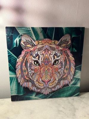 Färgglad tiger diamantmålning - Unik diamantmålning med ett detaljerat och färgglatt tigerhuvud i mandalastil. Bakgrunden har gröna blad och motivet är fullt av små pärlor i olika färger som skapar ett glittrigt och livligt intryck. Perfekt som dekoration för att ge rummet en cool vibe.