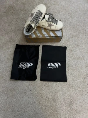 Golden Goose Superstar sneakers - Golden Goose Superstar sneakers i vit med grå stjärndetalj på sidan, silverfärgad häl och grå snörning. Skorna har en platt sula och rund tå. Materialet är skinn och mocka. Medföljer originalkartong och två svarta dustbags med GGDB-logga. Skorna är I storlek 41 och pris kan självklart diskuteras! 