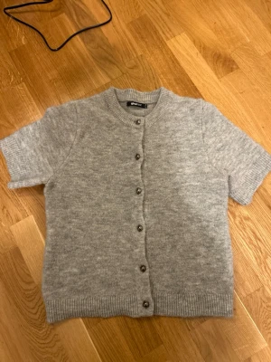 Grå stickad kofta från Gina Tricot - Mysig grå stickad kofta från Gina Tricot med korta ärmar och rund hals. Koftan har ribbade muddar och knäpps med silvriga knappar framtill. Perfekt att slänga över en t-shirt eller linne för en chill look.