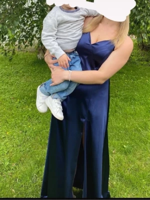 Marinblå balklänning - Säljer en marinblå långklänning med smala axelband och v-ringad hals. Klänningen har en slits framtill och är tillverkad i ett glansigt satinliknande materia. Använd en gång och är i fint skick!💙 tillsydd vid v-ringen. 