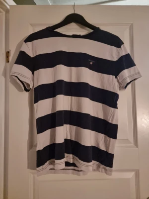 Randig t-shirt från GANT i bomull - Säljer en klassisk t-shirt från GANT med breda marinblå och vita ränder. T-shirten har korta ärmar, rund hals och broderad GANT-logga på bröstet. Materialet är mjuk bomull som känns skön mot huden. Perfekt för dig som gillar stilren och sportig look.