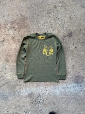 Olivgrön långärmad topp med tigertryck - Säljer en olivgrön långärmad topp från GUERRILLA med ett gult tigertryck på bröstet. Toppen har rund hals och är tillverkad i ett mjukt, ribbat material som sitter skönt mot kroppen. Perfekt för dig som gillar coola detaljer och streetwear.