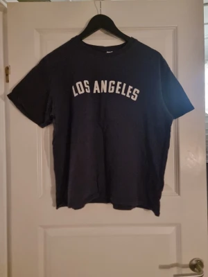 Svart t-shirt med Los Angeles-tryck - Svart t-shirt med vit text 'LOS ANGELES' tryckt på bröstet. Klassisk rund hals och korta ärmar. T-shirten är i mjuk bomull och har en avslappnad passform som passar perfekt till jeans eller shorts.