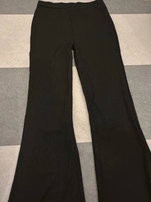 Svarta ribbade utsvängda byxor - Säljer ett par svarta byxor med ribbad struktur och utsvängda ben. De har en enkel design utan synliga fickor eller detaljer och är gjorda i ett mjukt, stretchigt material som sitter skönt. Perfekta för dig som gillar en chill och trendig look.