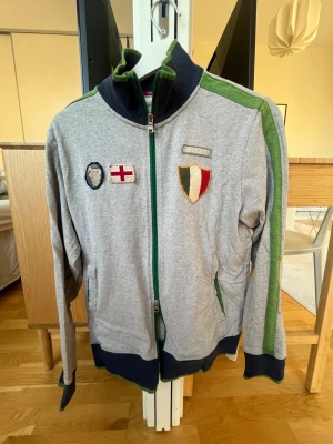 Grå ziptröja från Boggi Milano S - Grå ziptröja från Boggi Milano i storlek S med gröna och mörkblå detaljer. Tröjan har broderade märken på bröstet, gröna ränder längs ärmarna och mörkblå muddar. Hög krage och hel dragkedja framtill. Perfekt för en sportig och avslappnad stil.