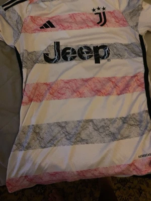 Juventus fotbollströja Adidas S - Juventus matchtröja från Adidas i storlek S. Tröjan har vita, grå och röda ränder med mönster, svart Juventus-logga och Jeep-tryck på bröstet. Tillverkad i lätt polyester med Aeroready-teknologi för bra ventilation. Perfekt för fotbollsfans!
