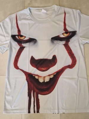 Vit t-shirt med Pennywise tryck - Säljer en vit t-shirt med ett stort, detaljerat tryck av Pennywise från filmen IT på både fram- och baksidan. T-shirten har korta ärmar och är tillverkad i ett mjukt syntetmaterial som känns skönt mot huden. Perfekt för dig som gillar skräck och vill sticka ut.