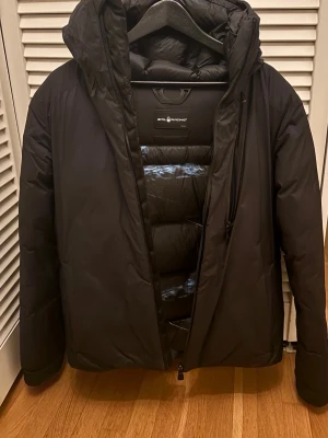 Svart dunjacka från Sail Racing - Svart dunjacka från Sail Racing med huva och dragkedja framtill. Jackan har en puffad design och är tillverkad i polyester med dunfyllning. Perfekt för kalla dagar och har en clean, modern look. Fickor med dragkedja och snygga detaljer på huvan.