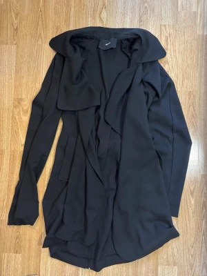 Svart trenchcoat från BikBok - Säljer en svart trenchcoat från BikBok i storlek S. Jackan har ett snyggt fall, bred krage och knytskärp i midjan för en clean look. Perfekt för dig som gillar stilrena och tidlösa plagg. Långa ärmar och mjukt material.