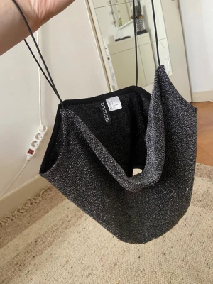 Glittrig svart bandeau topp Divided - Säljer en glittrig svart bandeau topp från Divided med tunna axelband. Toppen har en croppad passform och är tillverkad i ett syntetmaterial med silverglitter. Perfekt för att lyfta din outfit med lite extra shine.