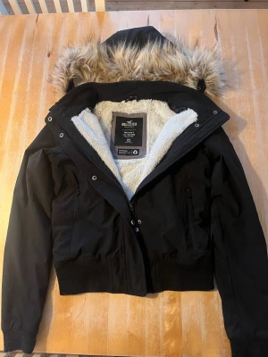 Svart bomberjacka från Hollister - Svart bomberjacka från Hollister med fluffigt vitt foder och huva med fuskpäls. Jackan har dragkedja framtill, två fickor med dragkedja och ribbade muddar. Perfekt för kalla dagar och har en snygg, kort passform. Jackan är änvänd fast har inga repor. Skriv för mer bilder eller några funderingar 💗💗