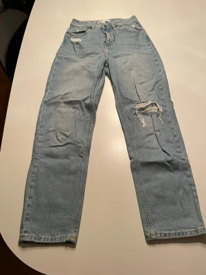 Ljusblå jeans med slitningar från H&M - Säljer ett par ljusblå jeans från H&M i storlek 34. De har raka ben, klassisk femficksdesign och snygga slitningar på låret och vid fickan. Jeansen  har en normal passform med midwaist. 