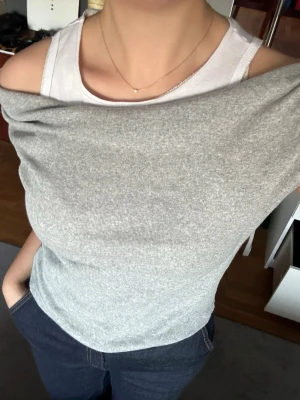 Grå omlott offshoulder topp från Zara - Säljer en unik grå omlott topp från Zara med offshoulder-design och vit undertopp. Toppen har en snygg draperad effekt över axeln och är ärmlös på ena sidan. Materialet är mjukt och stretchigt, perfekt för att skapa en cool och avslappnad look.