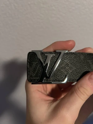  svart bälte med silvrig spänne - Säljer ett stilrent svart bälte  med klassiskt spänne i silver. Bältet har diskret monogrammönster och är tillverkat i präglat skinn. Perfekt accessoar för att lyfta din outfit med en lyxig touch.