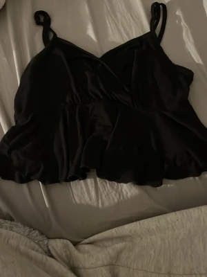 Svart omlott linnetopp med volang - Säljer en svart omlott linnetopp med dubbla smala axelband och volang nedtill. Toppen har en mjuk och glansig yta som ger en lyxig känsla. 