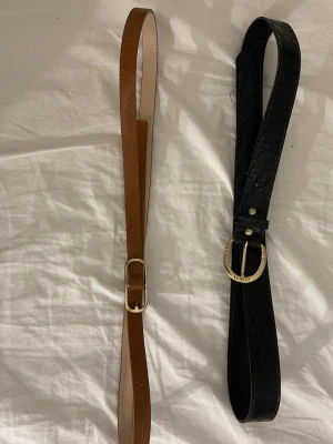 Svart och brun bälte med metallspänne - Två snygga bälten, ett i svart med krokodilpräglad yta och ett i klassisk brun färg. Båda har stilrena metallspännen i guldton. Det svarta bältet är bredare och har en cool textur, medan det bruna är smalare och har en slät finish. Perfekt för att lyfta din outfit.