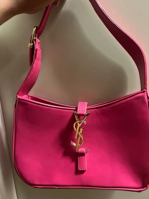 Rosa väska Yves Saint Laurent, YSL - Säljer en snygg rosa axelväska Yves Saint Laurent med guldigt YSL-emblem framtill. Väskan har en stilren, rektangulär form och är gjord i slätt skinn. Insidan har en dragkedja och en ljusrosa etikett med logga. Perfekt för dig som vill sticka ut med färg!