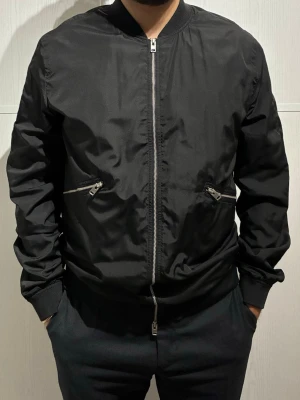 Svart tunn jacka J.Lindeberg - Svart jacka från J.Lindeberg med klassisk rund krage och silverfärgade dragkedjor framtill och vid fickorna. Jackan har långärmade muddar och är tillverkad i ett slätt syntetmaterial som ger en clean och modern look. Size Large men passar även till Medium.