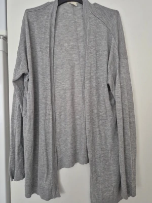 Kofta - Säljer en enkel och mjuk grå kofta från H&M i storlek S. Koftan har lång ärm, öppen framtill och är tillverkad i ett tunt stickat material. Perfekt att slänga över en t-shirt eller linne när det är lite kyligt ute.