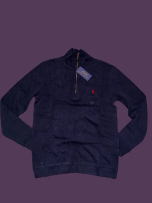 Marinblå half zip tröja Polo Ralph Lauren - Snygg marinblå tröja från Polo Ralph Lauren med half zip och ståkrage. Tröjan har ett rött broderat logomärke på bröstet och är tillverkad i mjuk bomull. Långärmad modell med normal passform, perfekt för chill höst- och vinterdagar.