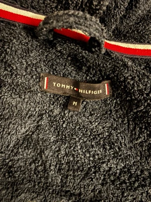 Mörkblå bummuls morgonrock Tommy Hilfiger - Säljer en mörkblå hoodie från Tommy Hilfiger i frottématerial, med huva och bälte i midjan. Tröjan har långa ärmar, fickor och broderad logga på ryggen. Perfekt för chill eller efter bad, med coola röda och vita detaljer på huvan.