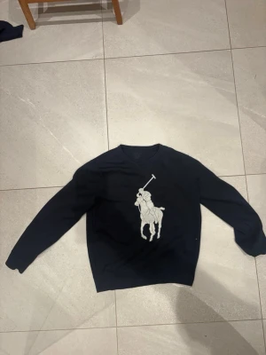 Mörkblå tröja från Ralph Lauren - Snygg mörkblå tröja från Ralph Lauren med stort broderat polospelare-motiv i vitt på bröstet. Tröjan har rund hals och långa ärmar, perfekt för dig som gillar klassisk och sportig stil.