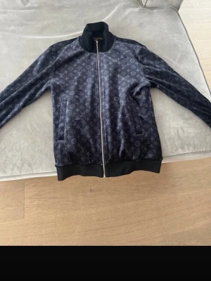 Louis Vuitton mörkblå sammetströja - Exklusiv mörkblå tröja från Louis Vuitton i mjuk sammet med ikoniskt monogrammönster över hela plagget. Tröjan har lång ärm, ribbade muddar, ståkrage och hel dragkedja med LV-detalj. Perfekt för dig som gillar lyxiga streetwear-vibes.