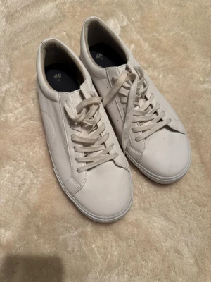 Vita sneakers från H&M, storlek 43 - Säljer ett par stilrena vita sneakers från H&M i storlek 43. Skorna har klassisk rund tå, platt sula och snörning framtill. Yttermaterialet är syntet med en slät finish och diskreta sömmar längs sidorna. Perfekta för dig som gillar minimalistisk och clean stil.