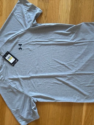 Grå tech t-shirt från Under Armour - Säljer en grå tech t-shirt från Under Armour i loose fit. T-shirten har korta ärmar, rund hals och är tillverkad i ett mjukt syntetmaterial som andas. Perfekt för dig som gillar sportig stil och vill ha något bekvämt till träningen. Helt oanvänd