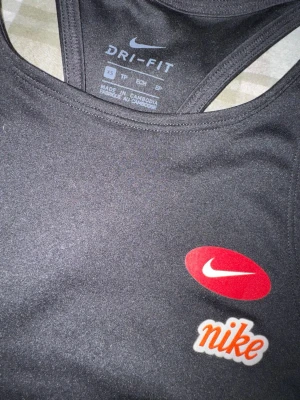 Svart träningstopp från Nike - Svart linnetopp från Nike med Dri-Fit-teknologi. Toppen har brottarrygg och tryckt Nike-logga i rött och vitt framtill. Perfekt för träning och sportiga aktiviteter.