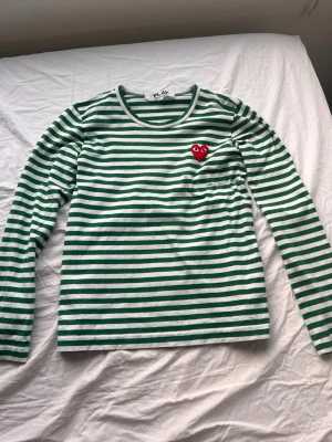 Randig långärmad topp Comme des Garçons - Grön och vit randig långärmad topp från Comme des Garçons PLAY med det ikoniska röda hjärtat broderat på bröstet. Tillverkad i mjuk bomull och har en klassisk rund halsringning. Perfekt för en stilren och trendig look.
