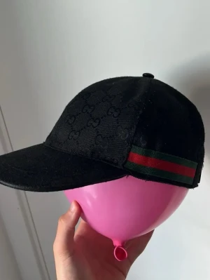 Keps - Säljer en svart  keps med diskret logomönster över hela tyget och klassiska gröna och röda ränder på sidan. Kepsen har justerbart spänne bak i skinn. Perfekt för dig som vill ha en snygg accessoar med lyxig känsla.