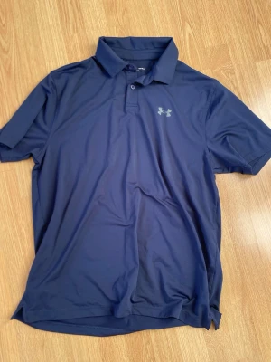 Marinblå polotopp från Under Armour - Säljer en marinblå polotopp från Under Armour i storlek L. Toppen har korta ärmar, klassisk krage och knappar framtill. Materialet är mjukt och syntetiskt, perfekt för träning eller sportiga dagar. Diskret logga på bröstet och loose passform.