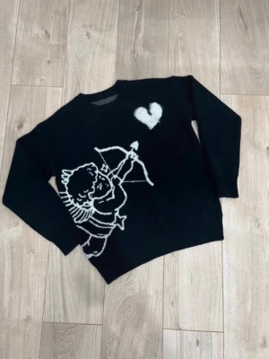 Svart stickad tröja med ängelmotiv - Svart stickad tröja från BRMG med vit illustration av en ängel som skjuter pil mot ett hjärta på framsidan. Baksidan har texten 'I WOULD RATHER NOT LOVE' och fler vita detaljer. Rund hals och långa ärmar. Perfekt för dig som gillar statement-plagg.