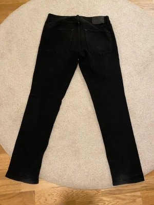 Svarta reguler jeans från Weekday - Säljer ett par svarta reguler jeans från Weekday med klassisk femficksdesign och knappgylf. Jeansen har smal passform och är tillverkade i stretchigt denim för extra komfort. Perfekta för dig som gillar en stilren och enkel look.