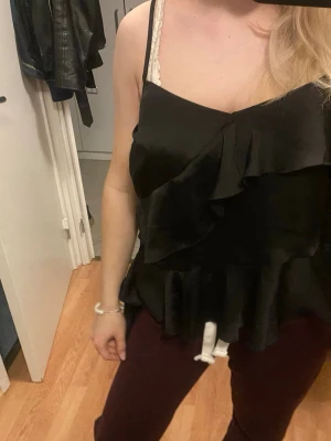 Svart omlott linnetopp med volang - Säljer en svart omlott linnetopp med volangdetaljer och smala axelband. Toppen har en glansig finish och är tillverkad i ett mjukt, lätt material som ger en snygg siluett. Perfekt att styla med jeans eller kjol för en trendig look.