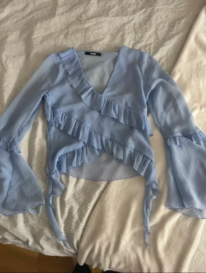 Ljusblå blus från Bik Bok - Supersöt ljusblå omlottblus från Bik Bok med v-ringning och breda ärmar. Blusen har volangdetaljer längs halsringningen och framtill, perfekt för vår och sommar. 