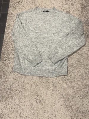 Grå stickad tröja från LMTD - Mysig grå stickad tröja från LMTD med rund hals och ribbade muddar vid ärmslut och nederkant. Perfekt för kyliga dagar och enkel att matcha med jeans. Tröjan har en klassisk och stilren look som passar till många olika outfits.