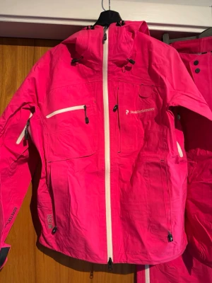 Peak Performance rosa vindjacka - Peak Performance vindjacka i stark rosa färg med vita dragkedjor och flera fickor. Jackan har huva, lång ärm och är tillverkad i slitstark Gore-Tex Pro Shell. Perfekt för dig som gillar att sticka ut och vill ha något funktionellt och snyggt.