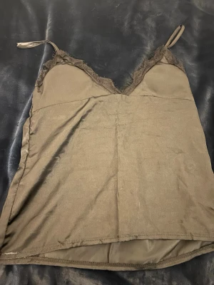 Svart  satin linnetopp med spets - Säljer en svart linnetopp i satin med tunna justerbara axelband och dekorativ spets längs v-ringad halsringning. Toppen har en glansig finish och är ärmlös, perfekt för att styla med jeans eller kjol. Spetsdetaljerna ger en extra touch av elegans.