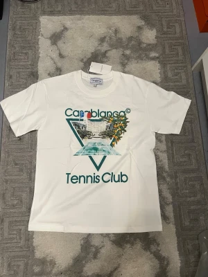 Vit Casablanca Tennis Club t-shirt - Casablanca Tennis Club t-shirt i vit bomull med coolt grafiskt tryck framtill som visar en pool, byggnad och apelsinträd. T-shirten har klassisk rund hals och korta ärmar. Perfekt för dig som gillar sportig och trendig stil.